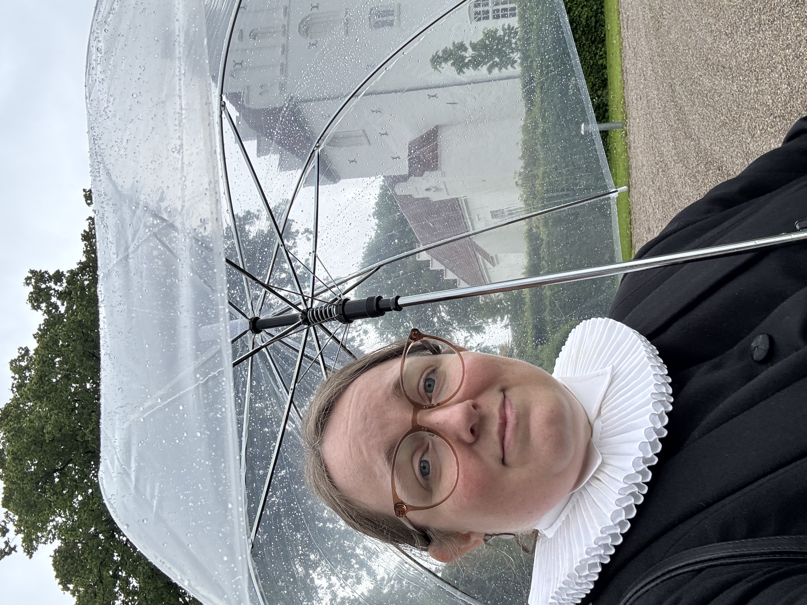 Sognepræst Eva Lykke Jensen under en gennemsigtig paraply i regnvejr og med Herringe Kirke i baggrunden.